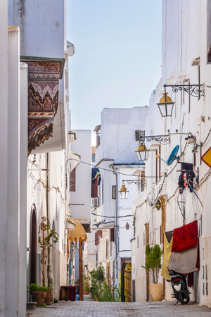 Rabat, Morocco â A quiet narrow alley in the historic medina of Rabat, lined with whitewashed buildings, traditional wooden doors, and decorative street lamps. The cobblestone street leads toward small local shops displaying colorful fabrics, creating an authentic atmosphere that reflects Moroccan architecture and daily life in the old city.の写真素材