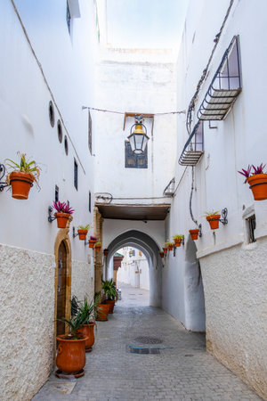 Rabat, Morocco â A quiet narrow alley in the historic medina of Rabat, lined with whitewashed buildings, traditional wooden doors, and decorative street lamps. The cobblestone street leads toward small local shops displaying colorful fabrics, creating an authentic atmosphere that reflects Moroccan architecture and daily life in the old city.の写真素材