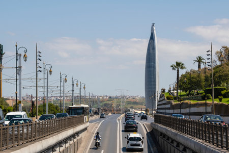 Rabat, Morocco â A modern view of Rabat's cityscape featuring the iconic Mohammed VI Tower, the tallest building in Africa. The sleek skyscraper rises above the urban skyline along a busy roadway with cars, buses, and palm-lined streets under a bright blue sky.の写真素材