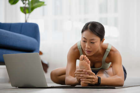 In the morning, an Asian woman is studying online yoga class, standing forward bend exercise, head...