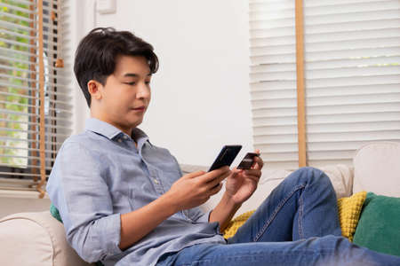 Asian man makes a payment via the internet. On the sofa at home, a shopper shops online using a credit card and a smartphone. The concept of internet shopping.の写真素材