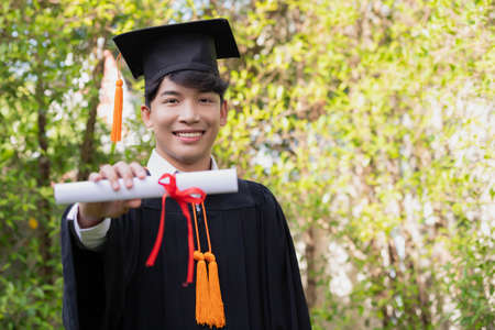 In the graden, a happy young Asian man wears graduation gowns and has a diploma.の写真素材