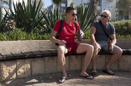 Two tourists of different ages drink beer and chat sitting on a benchの写真素材