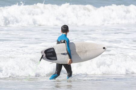 A teenager in a wetsuit with a surfboard in his hand walks in the water along the shore
