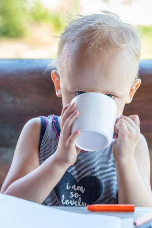 A baby with a cup of milk is sitting on the terrace of a wooden houseの写真素材