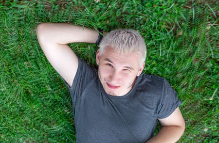 A cheerful guy in a gray T-shirt is lying on a green lawn with one eye squintedの写真素材