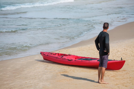 A man stands near a red kayak on a sandy beach.の写真素材