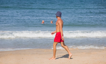 A muscular guy in red shorts and a blue panama hat walks along the seashoreの写真素材