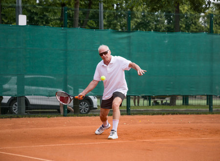 An elderly man playing tennis is hitting a forehand. The moving at camera. Open ground.の写真素材