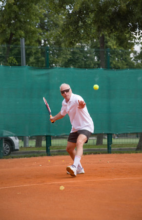 An elderly man playing tennis is hitting a forehand. The moving at camera. Open ground.の写真素材