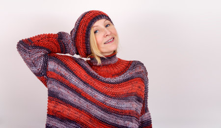A smiling woman, dressed in a knitted terracotta sweater and a beanie, has her hand behind her head....