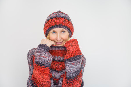 A woman dressed in a knitted terracotta sweater and a beanie pulls the collar of the sweater over...