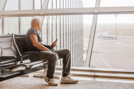 A mature man looks through a panoramic glass wall at the airfield, sitting in the departure hall of the airportの写真素材