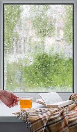 A womans hand with a cup of tea. An open book and a soft blanket. The window glass is covered with...