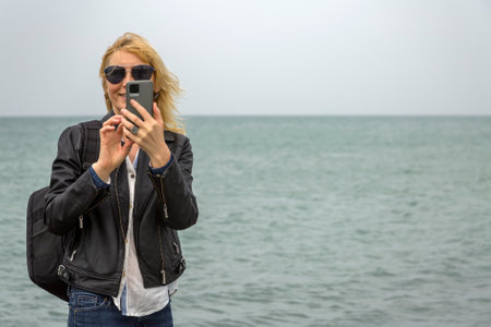 An adult woman in a black jacket and with a backpack on her shoulders takes photos on her smartphone, standing against the background of the sea.の写真素材