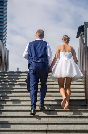 The tired bride and groom go up the stairs barefoot after a walk on a hot summer wedding day. Rear viewの写真素材