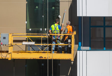 A construction winch with workers sealing joints between the facade panels of a modern building....