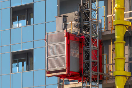 A red construction elevator rising along the facade of a modern building and a yellow plastic pipe for dumping construction trashの写真素材