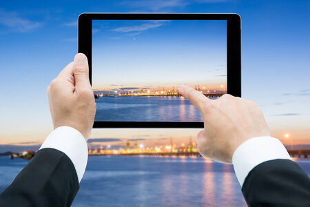 Businessman hands tablet taking pictures Commercial docks at sunset with ship and cranes