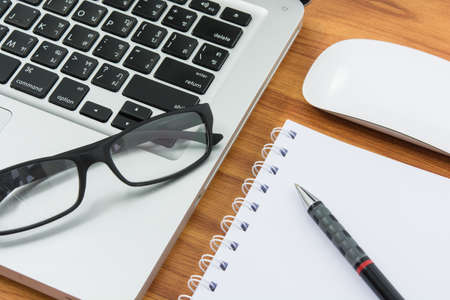 Blank business laptop, mouse, pen, note and glasses on wooden table