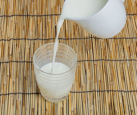 Milk from ceramic jug pouring into glass on wooden table backgroundの写真素材