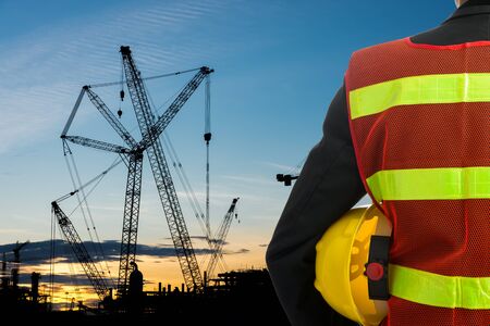 Hand or arm of engineer hold yellow plastic helmet in front of buildings and construction cranesの写真素材