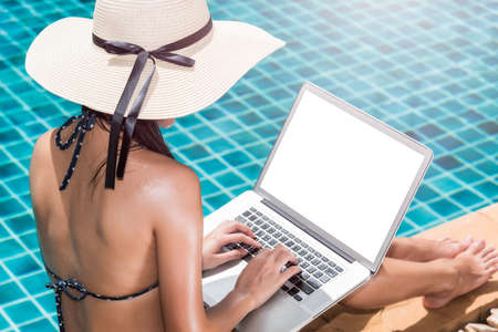 Asian Young woman in big hat relaxing on poolside with laptop computer blue water