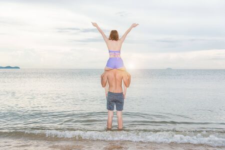 Young portrait couple having fun woman sitting on the shoulder show hands up at the beachの写真素材