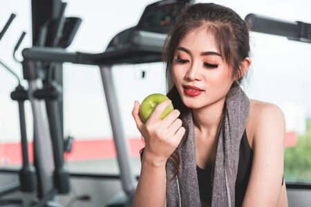 Young woman in sportswear holding green apple at fitness gym, Girl sitting on stepper taking breakの写真素材