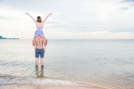Young portrait couple having fun woman sitting on the shoulder show hands up at the beachの写真素材
