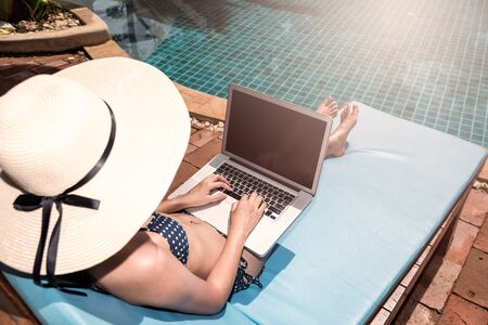 Beautiful Woman using laptop computer luxury relaxing sitting deck chair on resort near swimming pool blue waterの写真素材
