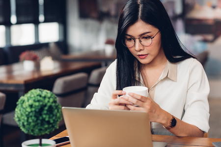 Freelancer woman holding coffee cup mug and using laptop computer on table in coffee cafe shopの写真素材