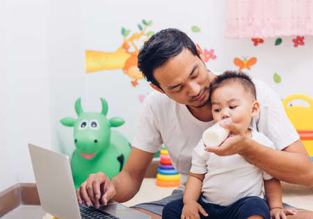Father acting Mom feeding milk his son baby 1 year old while working on laptop computer in the home.の写真素材