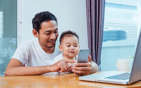 Man father working on laptop computer and using smartphone technology with son from homeの写真素材