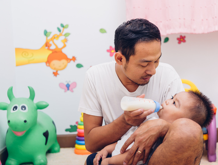Father acting Mom feeding milk his son baby 1 year old on chair in the houseの写真素材
