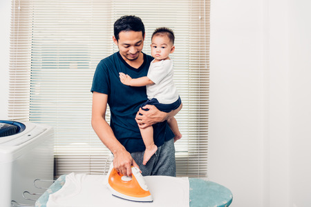 Man father ironing clothes son and he carry son in homeの写真素材