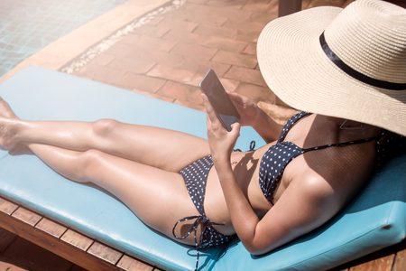 Beautiful girl woman with big hat happy use smartphone relaxing near swimming pool blue waterの写真素材