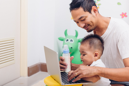 Father acting Mom feeding milk his son baby 1 year old while working on laptop computer in the home.の写真素材