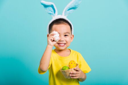 Asian cute little child boy smile beaming wearing bunny ears and a yellow T-shirt, standing to holds colored easter eggs instead of eyes on blue background with copy space for textの写真素材