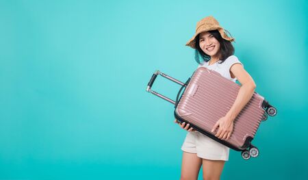 Portrait happy Asian beautiful young woman standing wear white t-shirt, holidays travel concept, her hugging suitcase bag and looking to camera, shoot photo in a studio on a blue backgroundの写真素材