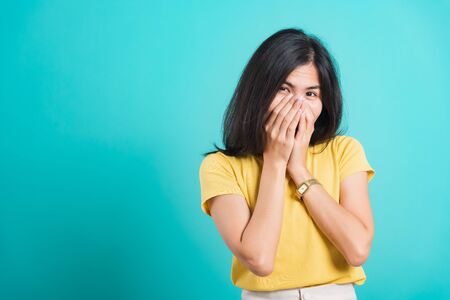 Portrait Asian beautiful happy young woman wears yellow t-shirt she happy excited laughs covering mouth with hands, shoot photo in a studio, on a blue background, with copy spaceの写真素材