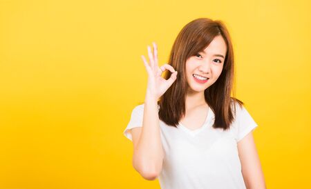 Asian happy portrait beautiful cute young woman teen standing wear t-shirt showing gesturing ok sign with fingers looking to camera isolated, studio shot on yellow background with copy space for textの写真素材