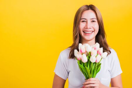 Portrait Asian Thai beautiful happy young woman smiling, screaming excited hold flowers tulips bouquet in hands and looking to camera, studio shot isolated on yellow background, with copy spaceの写真素材