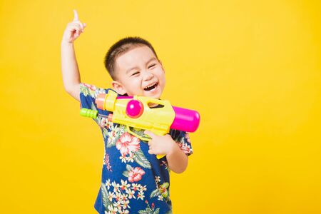 Portrait happy Asian cute little children boy smile standing so happy wearing flower shirt in Songkran festival day holding water gun, studio shot on yellow background with copy spaceの写真素材