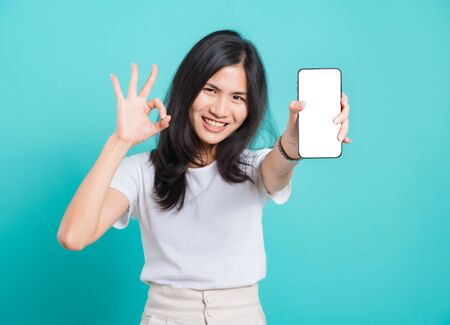 Portrait Asian beautiful happy young woman standing smile, holding mobile phone and showing ok gesture, shoot photo in studio focus phone on blue background, with copy spaceの写真素材
