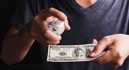 Closeup man wearing protective face mask applying sanitizer spray to 100 dollar money for cleaning,...