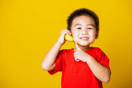 Happy portrait Asian child or kid cute little boy attractive smile wearing red t-shirt playing holds banana fruit pretending to be like a telephone, studio shot isolated on yellow backgroundの写真素材