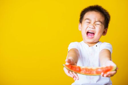 Happy portrait Asian child or kid cute little boy attractive laugh smile playing holds cut watermelon fresh for eating, studio shot isolated on yellow background, healthy food and summer conceptの写真素材