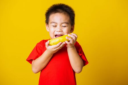 Happy portrait Asian child or kid cute little boy attractive smile wearing red t-shirt playing holds banana fruit, studio shot isolated on yellow backgroundの写真素材