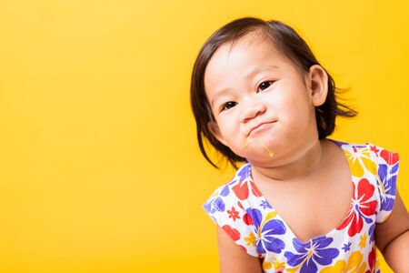 Happy portrait Asian baby or kid cute little girl attractive laugh smile wearing dick pattern shirt holds and eating sweet wooden ice cream, studio shot isolated on yellow background, summer conceptの写真素材